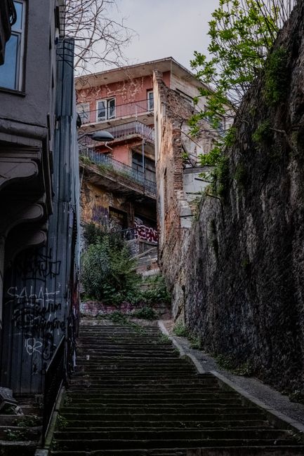 A narrow alleyway in an urban setting with a stone-paved surface leading to a short flight of stairs at the end, surrounded by closely packed multi-storey buildings with various window styles and balc