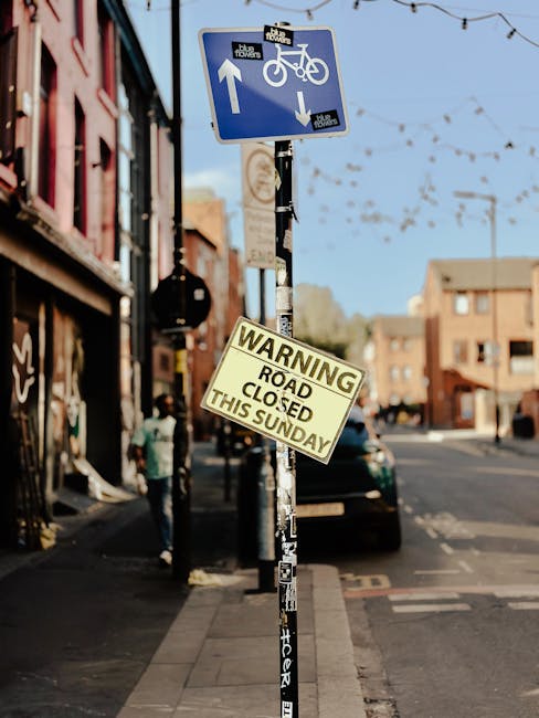 A street scene showing a metal pole with multiple signs attached, including a blue bicycle and pedestrian traffic sign indicating shared use of the path, and a yellow warning sign stating 'Road Closed